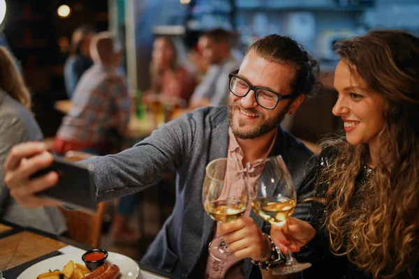 Linda pareja caucásica de moda sentada en el restaurante en la cena sosteniendo copas de vino y tomando selfie . —  Fotos de Stock