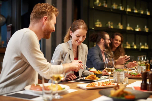 Симпатичный кавказский имбирь вытаскивает еду из тарелки своей девушки, сидя за ужином в ресторане. На заднем плане их друзья . — стоковое фото