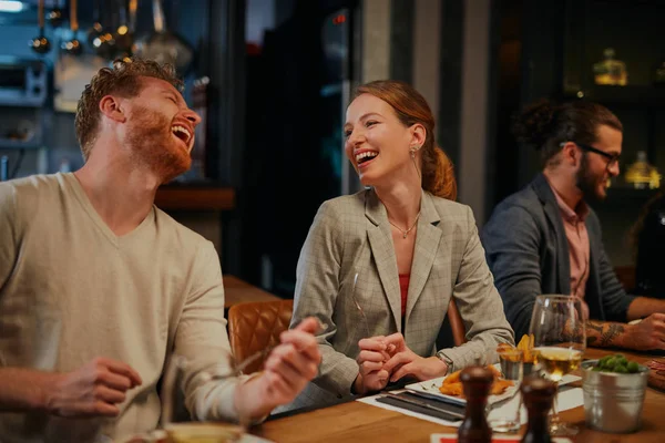 Charming smiling caucasian blonde sitting in restaurant, eating dinner and chatting with friend. In background are friends. — Stock Photo, Image