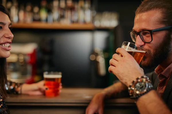 Junger gutaussehender kaukasischer bärtiger Mann mit Brille, der sich in der Kneipe an den Tresen lehnt, Bier trinkt und mit einem Freund plaudert. — Stockfoto