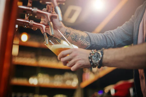 Tätowierter kaukasischer Barmann, der Bier einschenkt, während er in der Kneipe steht. Selektiver Fokus auf die Hand. — Stockfoto