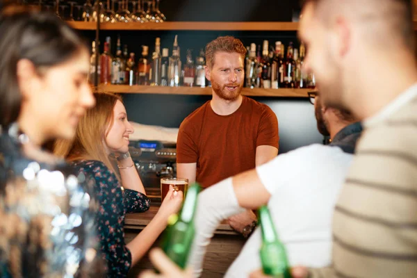Vrolijke vrienden leunend op een bar, bier drinkend en babbelend met de barman. Avond uit.. — Stockfoto
