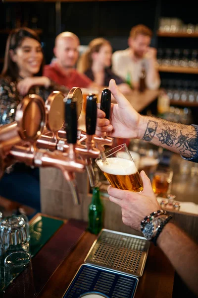 Cropped picture of tattooed barman pouring beer in glass. In background is group of friends sitting at counter. Pub interior. — Stock Photo, Image