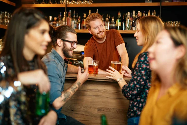 Amigos alegres apoyados en el mostrador del bar, bebiendo cerveza y charlando con el camarero. Noche fuera. . — Foto de Stock