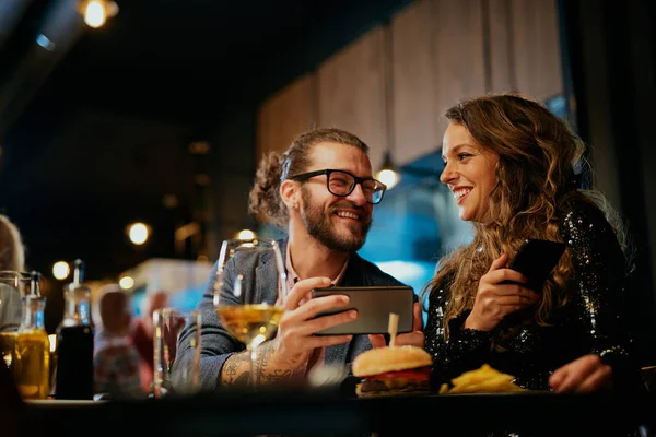 Charmant homme hipster caucasien prenant des photos de sa nourriture alors qu'il était assis au restaurant. À côté de lui est sa petite amie tenant un téléphone intelligent et le regardant . — Photo