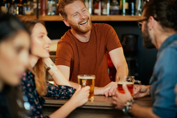 Χαρούμενοι φίλοι ακουμπάνε στον πάγκο του μπαρ, πίνουν μπύρα και κουβεντιάζουν με τον μπάρμαν. Νυχτερινή έξοδος. — Φωτογραφία Αρχείου
