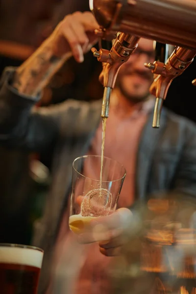 Tatuado barman caucasiano derramando cerveja enquanto estava de pé no pub. Foco seletivo na mão . — Fotografia de Stock