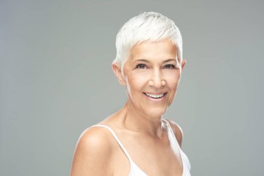 Gri arka plan önünde poz kısa gri saçlı güzel gülümseyen yaşlı kadın. Güzellik fotoğrafçılığı.