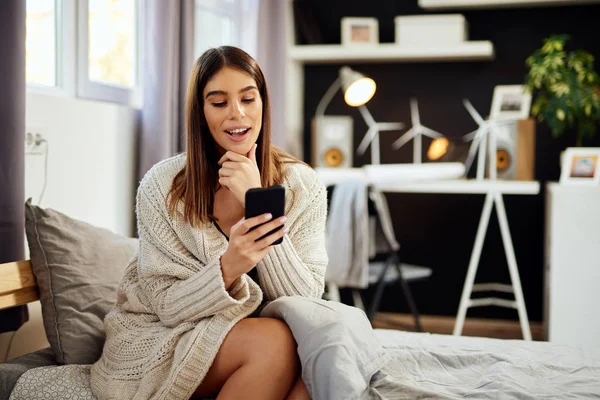 Hermosa morena caucásica sonriente sentada vestida con suéter beige sentada en la cama en el dormitorio y usando un teléfono inteligente . — Foto de Stock