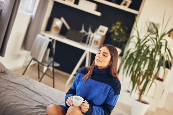 Charmante Brünette in blauem Pullover sitzt morgens im Schlafzimmer und trinkt Kaffee. — Stockfoto