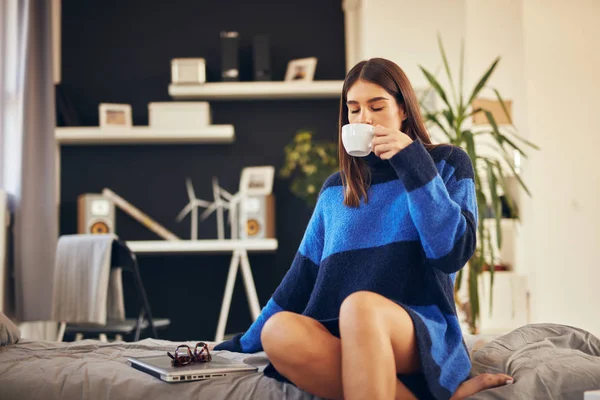 Charming brunette in blue sweater sitting in bedroom in morning and drinking coffee. — Stock Photo, Image