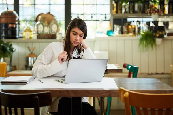 Mooie Kaukasische elegante zakenvrouw zitten in cafe, het houden van bril en met behulp van laptop. — Stockfoto