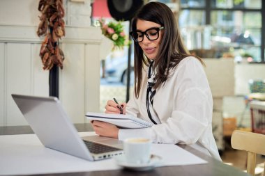 Kafeteryada oturan ve defterine görevler yazan beyaz tenli bir iş kadını. Masanın önünde dizüstü bilgisayar var..