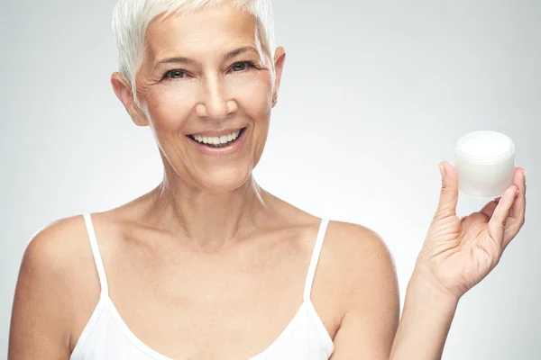 Prachtige glimlachend blanke Senior vrouw uitproberen van nieuwe anti-age crème en kijken naar de camera. Beauty fotografie. — Stockfoto