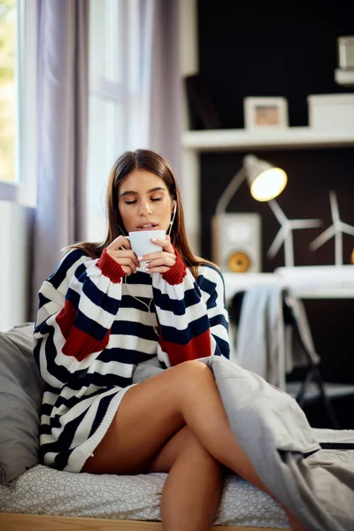 Hermosa morena caucásica sonriente vestida con suéter sentada en la cama en el dormitorio, usando tableta y tomando café por la mañana . — Foto de Stock