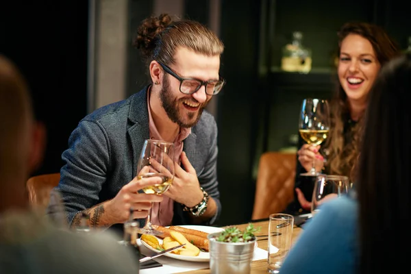 Handsome caucasian bearded hipster man with eyeglasses drinking wine and laughing while sitting with his friends in restaurant. — Stock Photo, Image