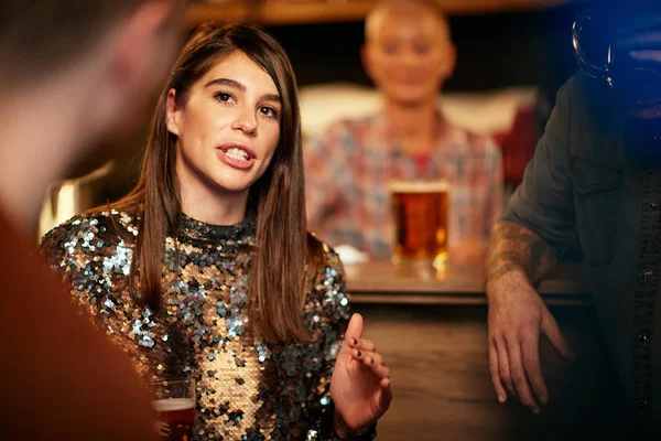 Όμορφη καυκάσια χαμογελαστή κομψή γυναίκα κρατώντας μπύρα και κουβεντιάζοντας με φίλους. Στο παρασκήνιο είναι μετρητής μπαρ. Pub εσωτερικό. — Φωτογραφία Αρχείου