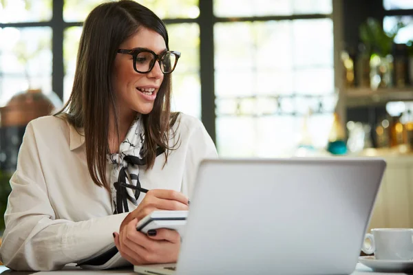 Encantadora mujer de negocios caucásica con gafas sentadas en la cafetería y tareas de escritura en el cuaderno. Delante de ella en la mesa está el ordenador portátil . — Foto de Stock