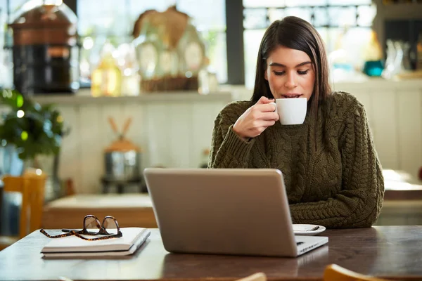 Attraktive kaukasische Brünette im Pullover sitzt im Café und trinkt Kaffee. Vor ihr liegt ein Laptop. — Stockfoto