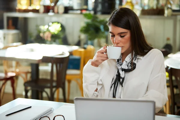 Aantrekkelijke Kaukasische elegante brunette zit in cafe, het drinken van koffie. Op tafel ligt een laptop. — Stockfoto