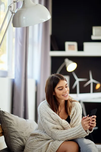 Atractiva morena caucásica vestida con suéter beige sentada en la cama en el dormitorio y usando un teléfono inteligente . — Foto de Stock