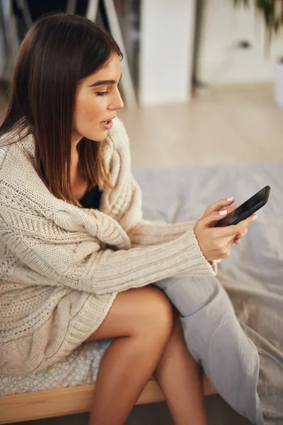 Vonzó kaukázusi barna ruhás bézs pulóver ül az ágyon a hálószobában, és használ okos telefon. — Stock Fotó
