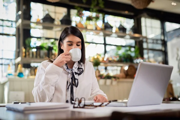 De jonge blanke zakenvrouw zit in een café, neemt een pauze van haar werk en drinkt verse koffie. Op tafel liggen laptop en papierwerk. — Stockfoto