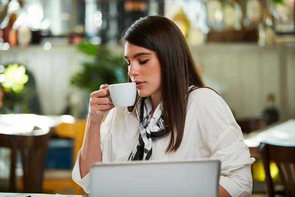 De jonge blanke zakenvrouw zit in een café, neemt een pauze van haar werk en drinkt verse koffie. Op tafel liggen laptop en papierwerk. — Stockfoto