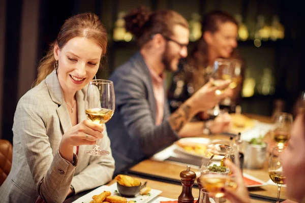 Hermosa morena caucásica vestida elegante posando con copa de vino mientras está sentada en el restaurante con sus amigos . —  Fotos de Stock