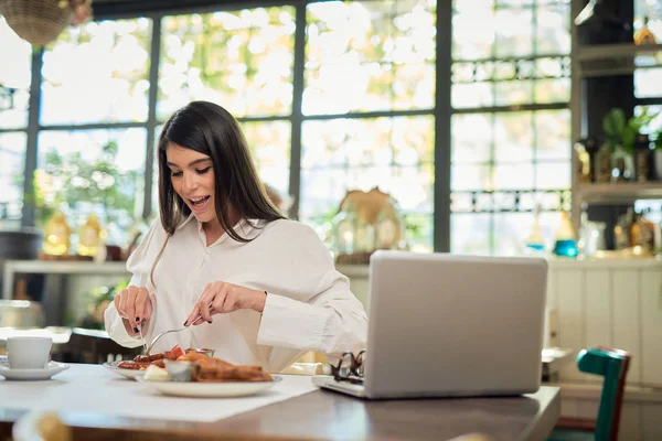 Prachtige Kaukasische elegante brunette zittend in restaurant en worstje etend als ontbijt. Op tafel ligt een laptop. — Stockfoto