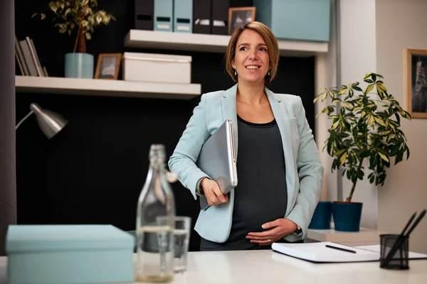 Positivt leende kaukasisk gravid affärskvinna som håller laptop under armhålan, vidrör magen och står på kontoret. Hon förbereder sig för ett affärsmöte med kollegor. — Stockfoto