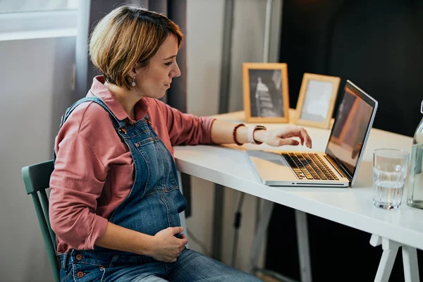 Attraktive kaukasische schwangere Frau sitzt zu Hause im Büro, berührt Bauch und nutzt Laptop für die Arbeit. — Stockfoto