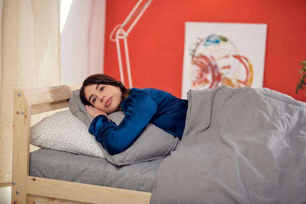 Atractiva morena caucásica vestida con pijamas azules acostada en la cama sobre el estómago. Hora de la mañana . — Foto de Stock