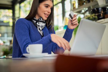 Kafede oturan, elinde kredi kartı olan ve bilgisayarını online alışveriş için kullanan çekici, beyaz tenli esmer. Laptopun yanındaki masada kahve var..