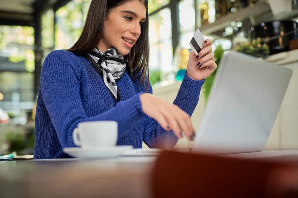 Attractive smiling caucasian brunette sitting in cafe, holding credit card and using laptop for online shopping. On table next to laptop is coffee.