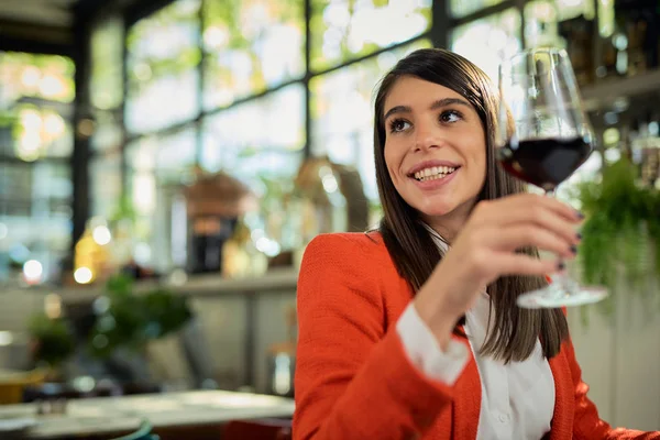Gyönyörű mosolygós kaukázusi barna ruhás elegáns ül az étteremben ebédszünetben, és kezében pohár vörösbor. — Stock Fotó