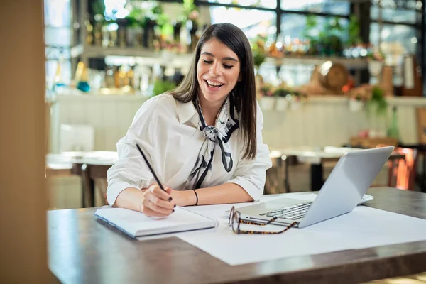 Pozitivní atraktivní mladá podnikatelka ve třicátých letech oblečená v košili sedí v kavárně a psaní úkolů v agendě. Na stole je laptop. Vzdálený obchodní koncept. — Stock fotografie