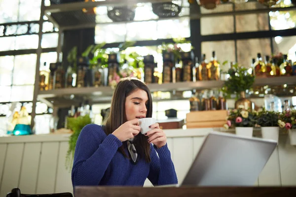 Atractiva morena caucásica en suéter azul sentada en la cafetería y disfrutando de café fresco. En la mesa está el portátil . —  Fotos de Stock