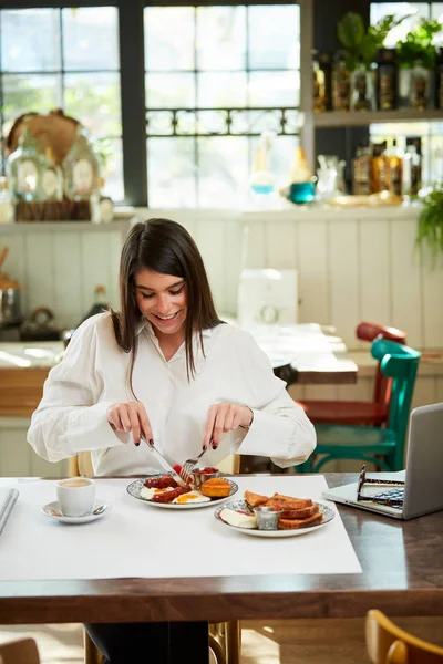 Charmante Kaukasische glimlachende brunette in shirt zittend in restaurant en het hebben van traditioneel ontbijt. Ze snijdt worstjes.. — Stockfoto