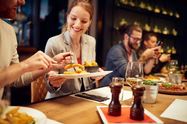 Happy caucasian couple sitting in restaurant and sharing food. In background are their friends using smart phones. — Stock Photo, Image