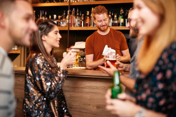 Un piccolo gruppo di migliori amici in piedi in un pub, bere birra, chiacchierare e divertirsi. Concentrati sul barista che pulisce il bicchiere. Vita notturna . — Foto Stock