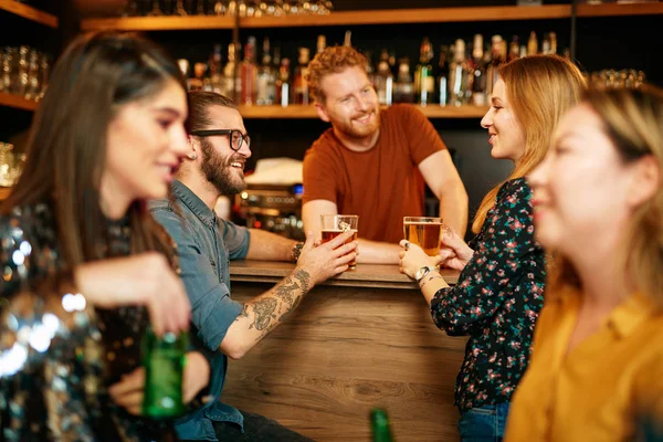 Pequeño grupo de amigos multiculturales de pie en el pub, apoyado en el mostrador del bar, charlando y sosteniendo pintas de cerveza. En primer plano están las chicas. Concéntrate en pareja cerca del mostrador . — Foto de Stock