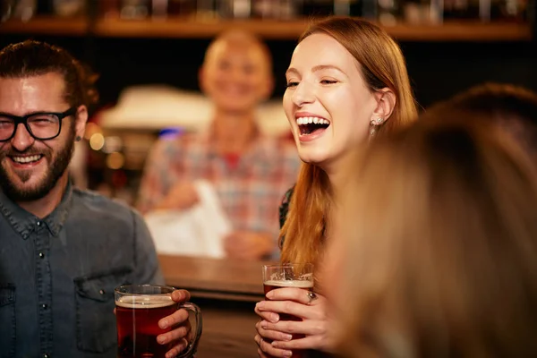 Wunderschöne kaukasische Frau lacht und hält ein Pint Bier in der Hand, während sie in der Kneipe steht, umgeben von ihren besten Freunden. — Stockfoto