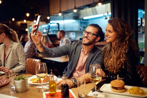 Schattig Kaukasisch glimlachend koppel verliefd zittend in restaurant en selfie nemend. Om hen heen zijn hun vrienden aan het chatten en dineren.. — Stockfoto