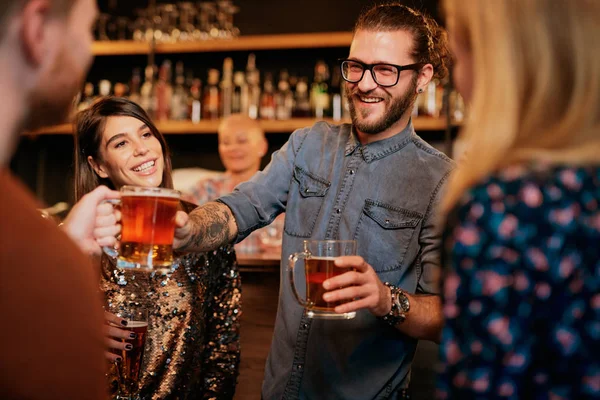 Homem hipster sorrindo em pé no pub com amigos e passando uma cerveja para seu amigo. Grupo de amigos se divertindo no pub. Vida noturna . — Fotografia de Stock