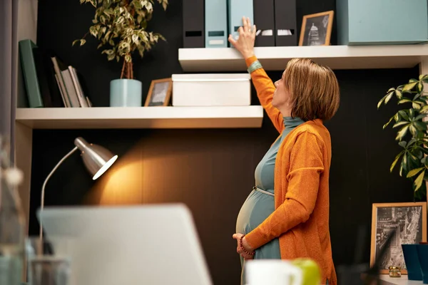 Sidovy av vacker gravid affärskvinna stående i ämbetet, vidrör magen och försöker nå pärm med viktiga dokument. — Stockfoto