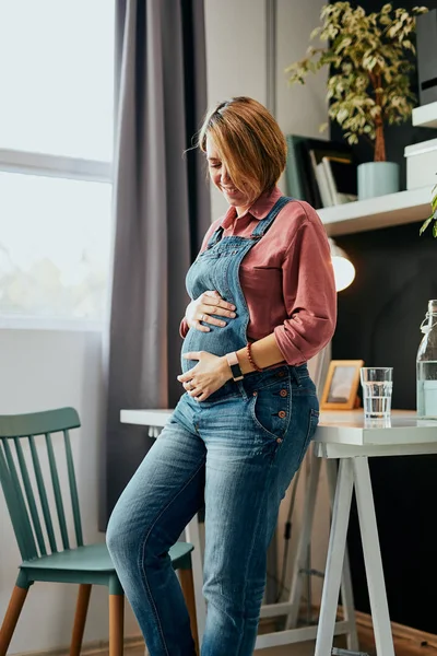 매력적 인 백인 장래 의임 신 한 임신부 가집 사무실에서 책상에 기대어 배에 손을 대는 모습. — 스톡 사진