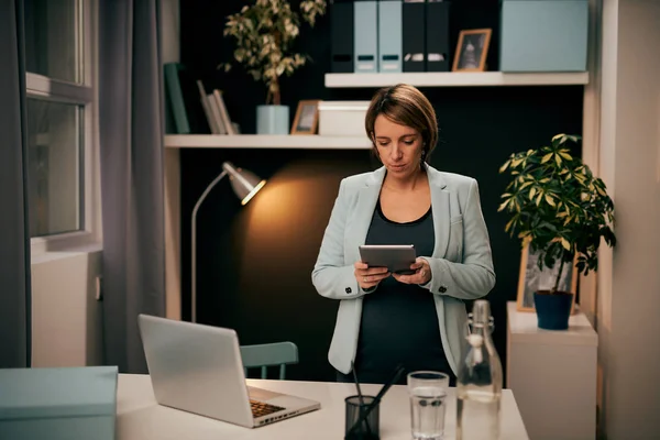 Takım elbiseli, beyaz tenli hamile bir iş kadını gece ofiste duruyor ve iş için tablet kullanıyor. İşkolik. Gece konseptinde çalışmak. — Stok fotoğraf