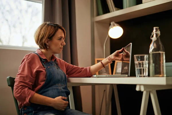 Γοητευτική καυκάσιος έγκυος επιχειρηματίας ντυμένος casual κάθεται στο γραφείο στο σπίτι, αγγίζοντας την κοιλιά και το άνοιγμα του φορητού υπολογιστή. Ετοιμάζεται για απομακρυσμένη δουλειά.. — Φωτογραφία Αρχείου