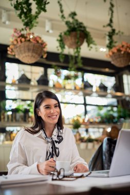 Kafeteryada oturan beyaz bir esmer, elinde bir fincan kahveyle işe ara veriyor. Masada dizüstü bilgisayar var.
